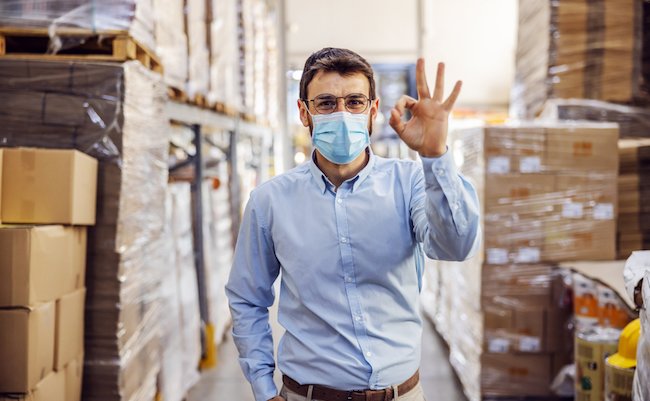 Young businessman with sterile protective mask on standing in warehouse and showing okay sign. Protection from corona virus/ covid-19.