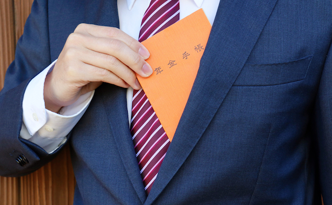 A man in a suit. An orange notebook with Japanese writing. Translation: Pension handbook.