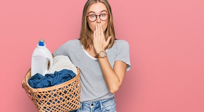 Beautiful blonde woman holding laundry basket and detergent bottle covering mouth with hand, shocked and afraid for mistake. surprised expression