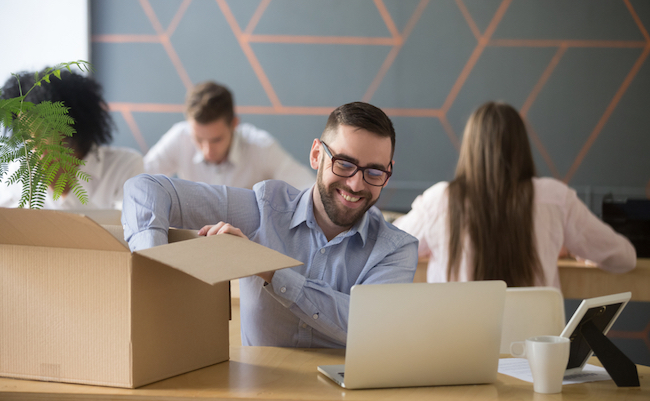 Smiling new male employee unpacking box with belongings at workplace, happy hired office worker newcomer on first working day concept, excited millennial businessman put laptop on desk in coworking