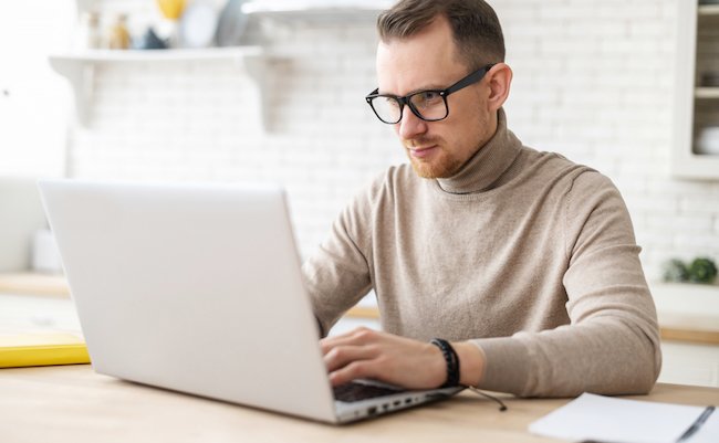 Serious focused determined young bearded businessman or freelancer in glasses and casual clothes sitting at kitchen table, typing on laptop, working on project, researching, writing email to client