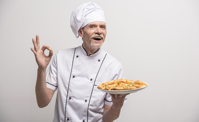 Senior male chief cook in uniform gesturing okay sign  and holding pizza on grey background.