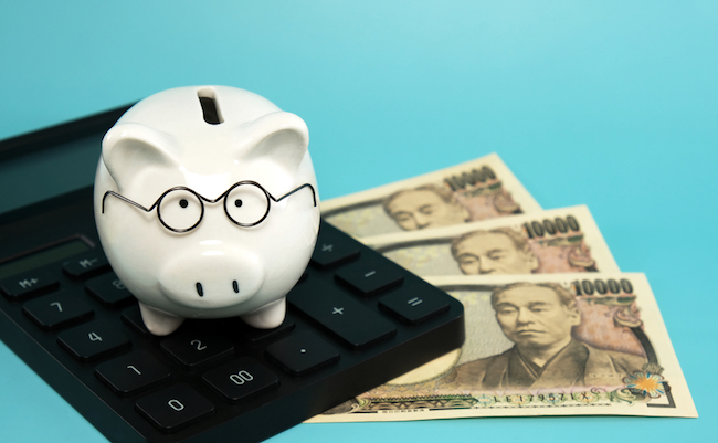 White piggy bank wearing reading glasses standing on calculator with Japanese yen banknotes on blue teal background. Concept for money savings plan for retirement, aged society or financial accounting