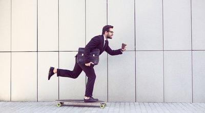 Toned image of businessman in black business suit on longboard walking with cup of coffee in his hand. Freelance man hurrying to his office.