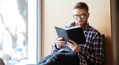 Young male student sitting on windowsill in library and reading book