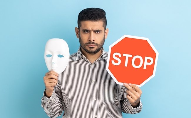 Portrait of serious businessman holding white mask with unknown face and red traffic sign, looking at camera, wearing striped shirt. Indoor studio shot isolated on blue background.