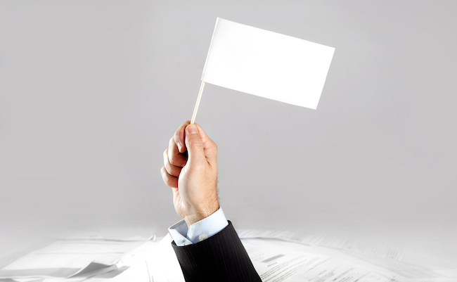 Hand of caucasian businessman emerging from office desk loaded of paperwork , invoices and a lot of papers and documents holding white flag overworked in giving up concept