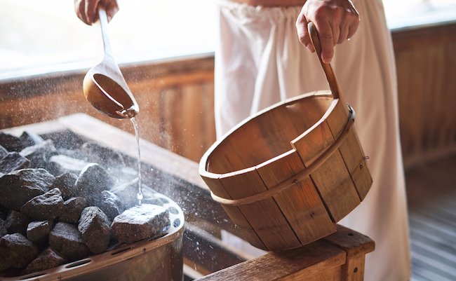 Man pouring water onto hot stone in sauna room. Steam on the stones, spa and wellness concept, relax in hot finnish sauna.