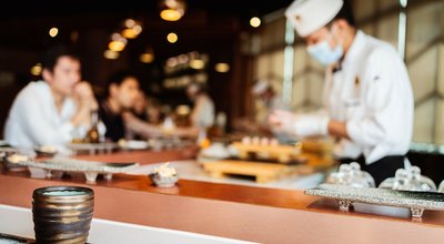 Japanese Omakase Restaurant counter focuses on Japanese ceramic tea cup with Blur chef cooking at the kitchen counter and directly serve to customers in the background.