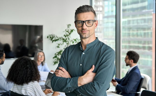 Smiling confident mature businessman leader looking at camera standing in office at team meeting. Male corporate leader ceo executive manager wearing glasses posing for business portrait arms folded.