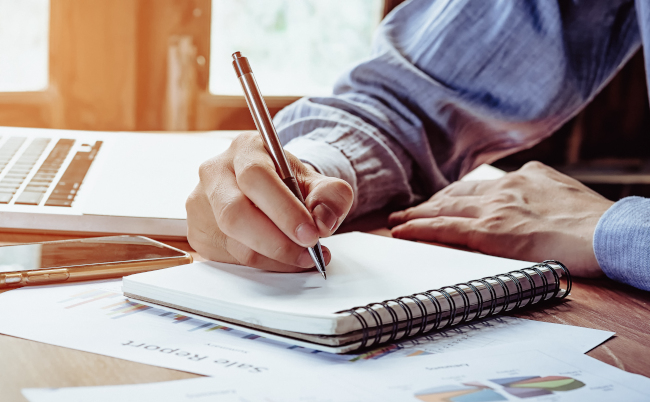 Businessman,Hands,With,Pen,Writing,Notebook,On,Office,Desk,Table