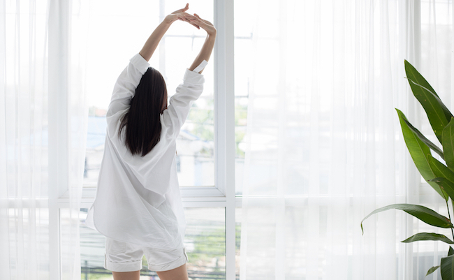 Asian woman waking up in her bed fully rested and open the curtains in the morning to get fresh air.