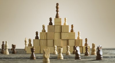 Business hierarchy; ranking and strategy concept with chess pieces standing on a pyramid of wooden building blocks with the king at the top with copy space.