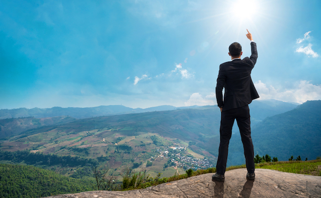 Back of successful businessman pointing up with his finger standing on top of mountain with sunlight sky background. Young man reaching goal, success and achievement in his life. Copy space.