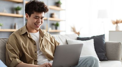 Portrait Of Smiling Asian Man Sitting On The Couch Working On Pc Laptop Indoors In Living Room. Happy Male Using Computer For Rest, Study Or Education, Typing On Keyboard, Surfing Internet