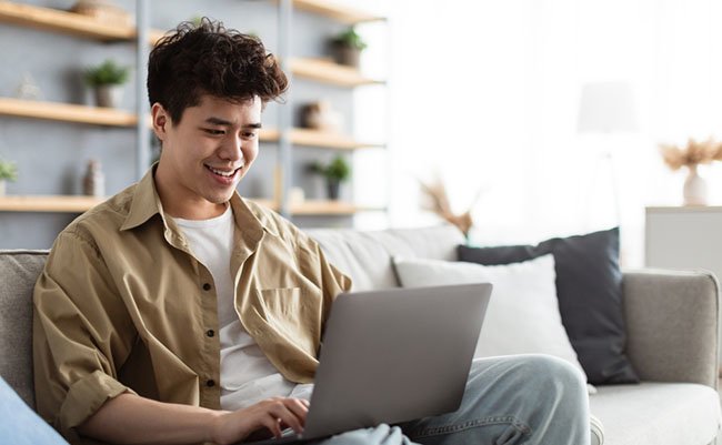 Portrait Of Smiling Asian Man Sitting On The Couch Working On Pc Laptop Indoors In Living Room. Happy Male Using Computer For Rest, Study Or Education, Typing On Keyboard, Surfing Internet