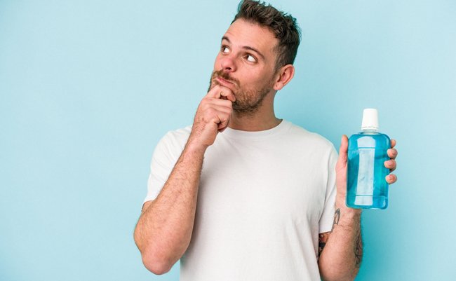 Young caucasian man holding mouthwash isolated on blue background looking sideways with doubtful and skeptical expression.