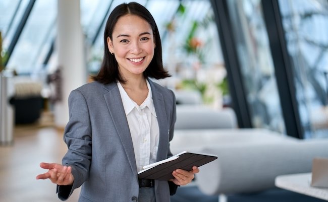 Happy young Asian saleswoman looking at camera welcoming client. Smiling woman executive manager, secretary offering professional business services holding digital tablet standing in office. Portrait