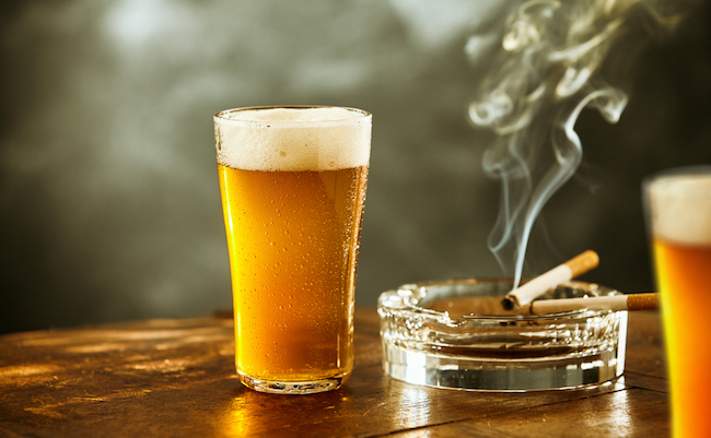 Ice cold lager or beer with a burning cigarette resting on a glass ashtray on a rustic wooden counter in a tavern, nightclub or pub, low angle close up