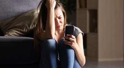 Front view of a sad teen checking phone sitting on the floor in the living room at home with a dark background