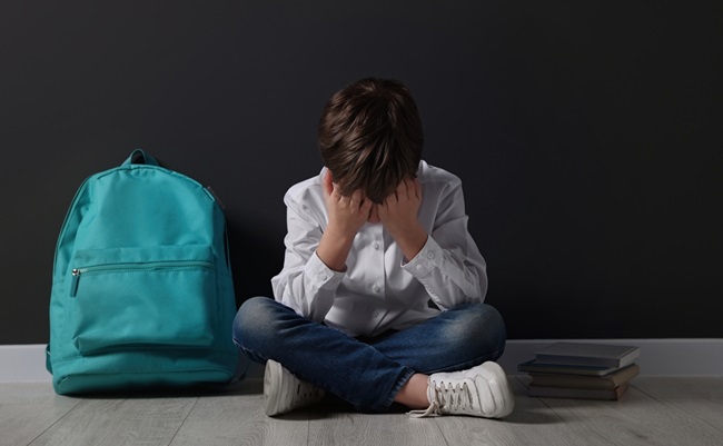 Upset,Boy,With,Backpack,Sitting,On,Floor,Near,Black,Wall.