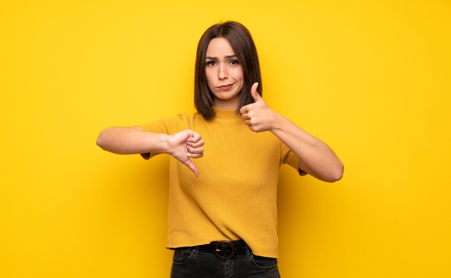 Young,Woman,Over,Yellow,Wall,Making,Good-bad,Sign.,Undecided,Between