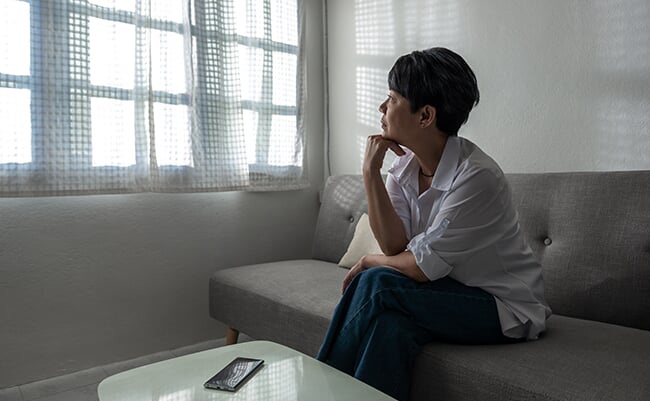 A senior gray-haired Asian woman is sitting on a couch at her home. She alone in the dark and waiting for a call, an unhappy elder woman lost in thoughts missing remembering past, feeling lonely.
