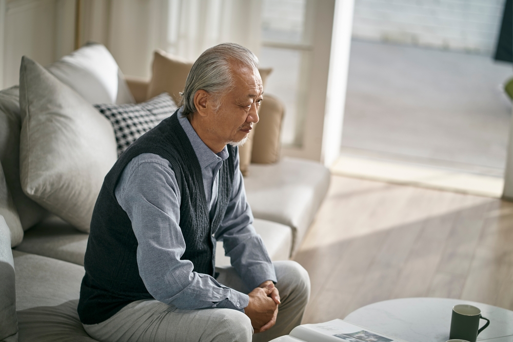 Sad,Asian,Senior,Man,Sitting,On,Couch,In,Living,Room
